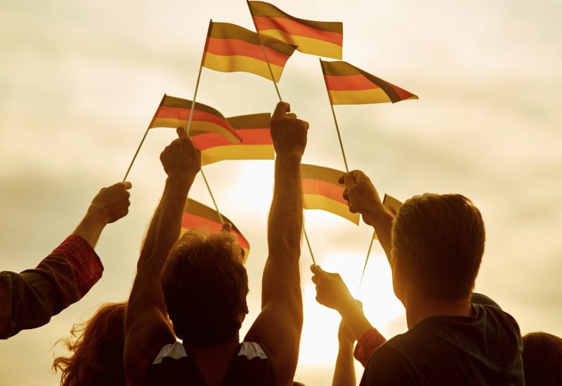 People hold german flags. Silhouette of patriotic deutsch crowd, rear view. Gloving evening sun background.